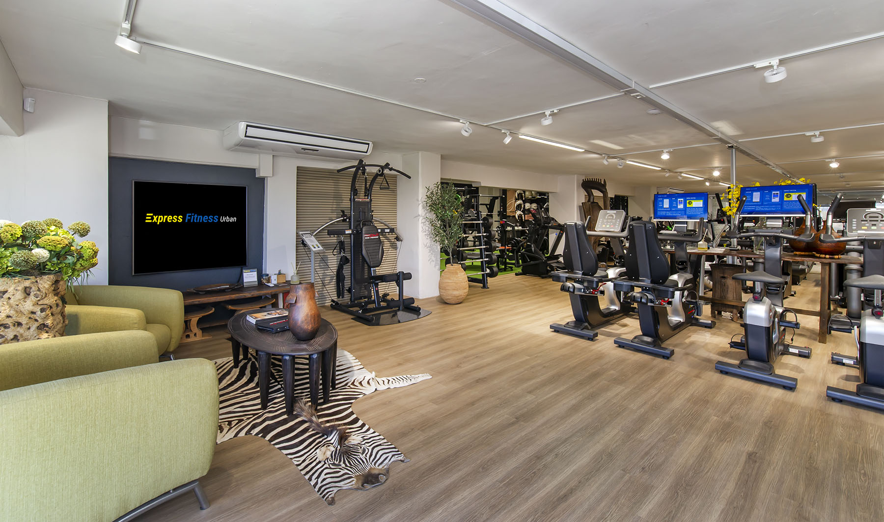 A wide view of the Express Fitness Urban showroom. In the foreground is a lounge area with green armchairs and a zebra-print rug, and in the background is a large display of cardio machines including recumbent and upright bikes.