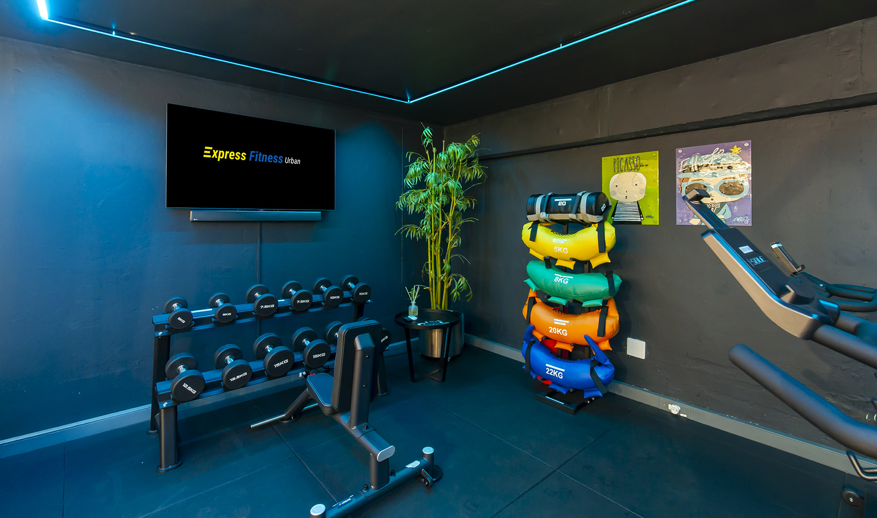 A compact section of the Express Fitness Urban showroom with dark walls and blue neon lights. The space features a branded TV screen, a dumbbell rack with an adjustable bench, and a stack of multi-coloured functional training bags.