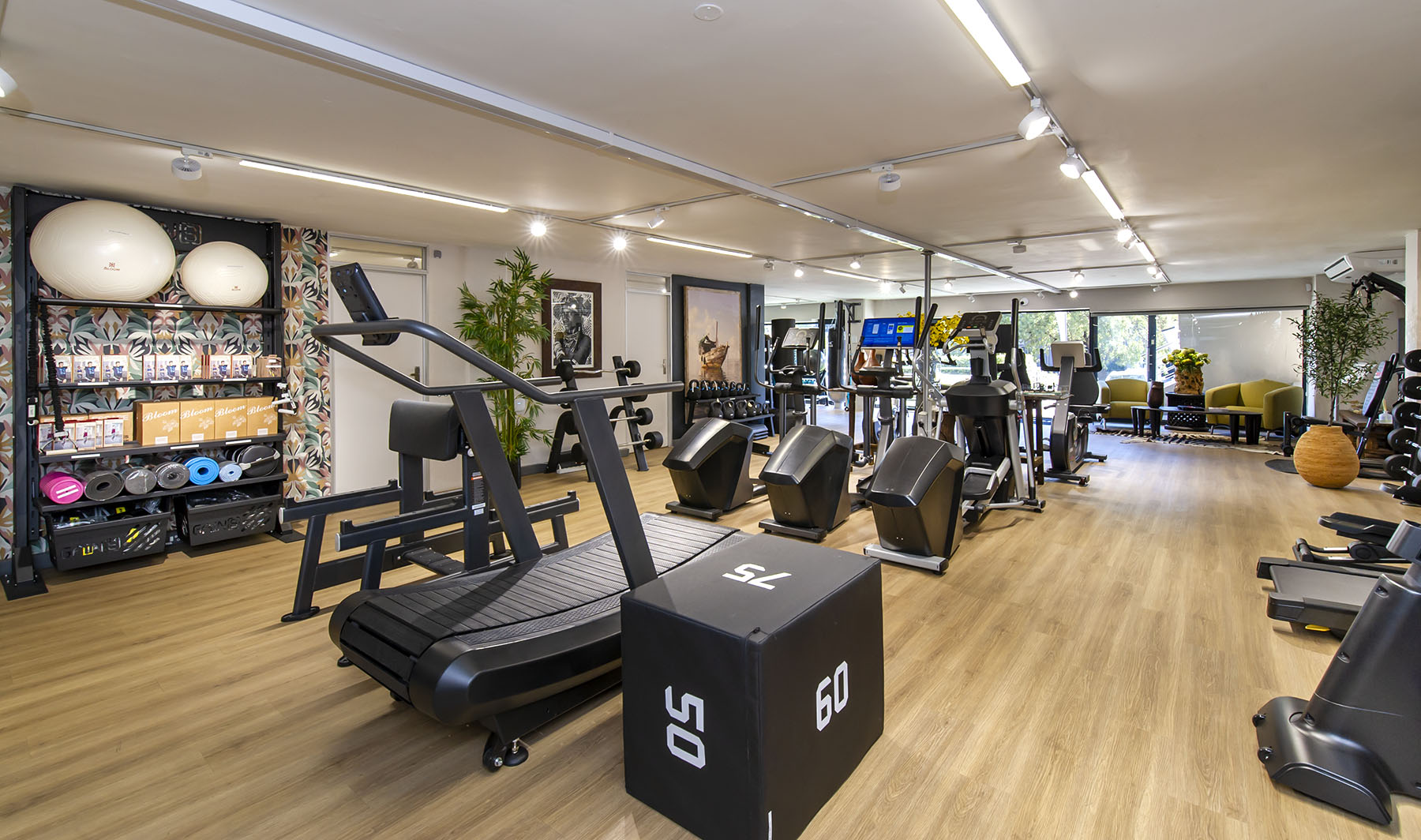 A wide-angle view of the Express Fitness Urban showroom in Sandton, Gauteng, showcasing a non-motorised treadmill and a plyometric box in the foreground. A shelving unit with fitness accessories is on the left, and a range of cardio machines including ellipticals and treadmills are visible in the background, alongside a lounge area.